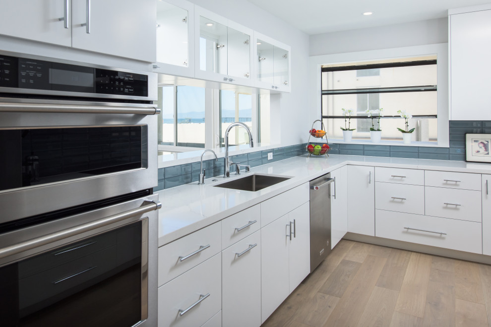 Photo of a small nautical u-shaped enclosed kitchen in Los Angeles with a single-bowl sink, flat-panel cabinets, white cabinets, engineered stone countertops, blue splashback, glass tiled splashback, stainless steel appliances, medium hardwood flooring, no island, brown floors and white worktops.