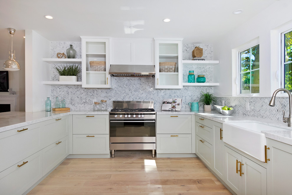 Photo of a coastal u-shaped kitchen in Los Angeles with a belfast sink, shaker cabinets, grey cabinets, grey splashback, stainless steel appliances, light hardwood flooring and a breakfast bar.