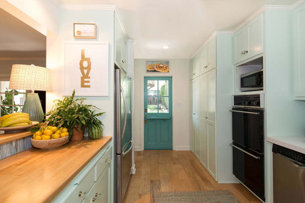 Example of a mid-sized beach style l-shaped medium tone wood floor enclosed kitchen design in Los Angeles with a peninsula, shaker cabinets, green cabinets, solid surface countertops, green backsplash, glass sheet backsplash, stainless steel appliances and a single-bowl sink