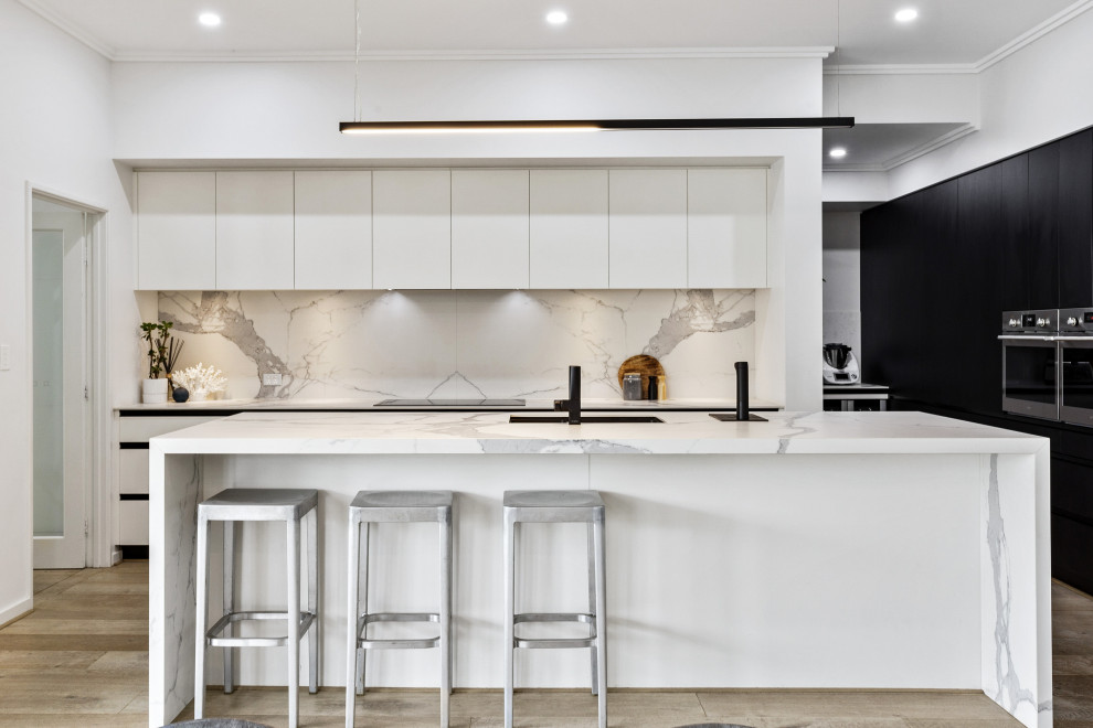 Bild på ett funkis vit vitt l-kök, med en undermonterad diskho, släta luckor, vita skåp, vitt stänkskydd, stänkskydd i sten, rostfria vitvaror, mellanmörkt trägolv, en köksö och brunt golv