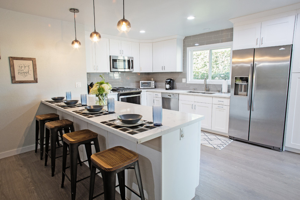 Photo of a medium sized contemporary u-shaped kitchen/diner in Orange County with a submerged sink, shaker cabinets, white cabinets, engineered stone countertops, grey splashback, porcelain splashback, stainless steel appliances, laminate floors, a breakfast bar and grey floors.