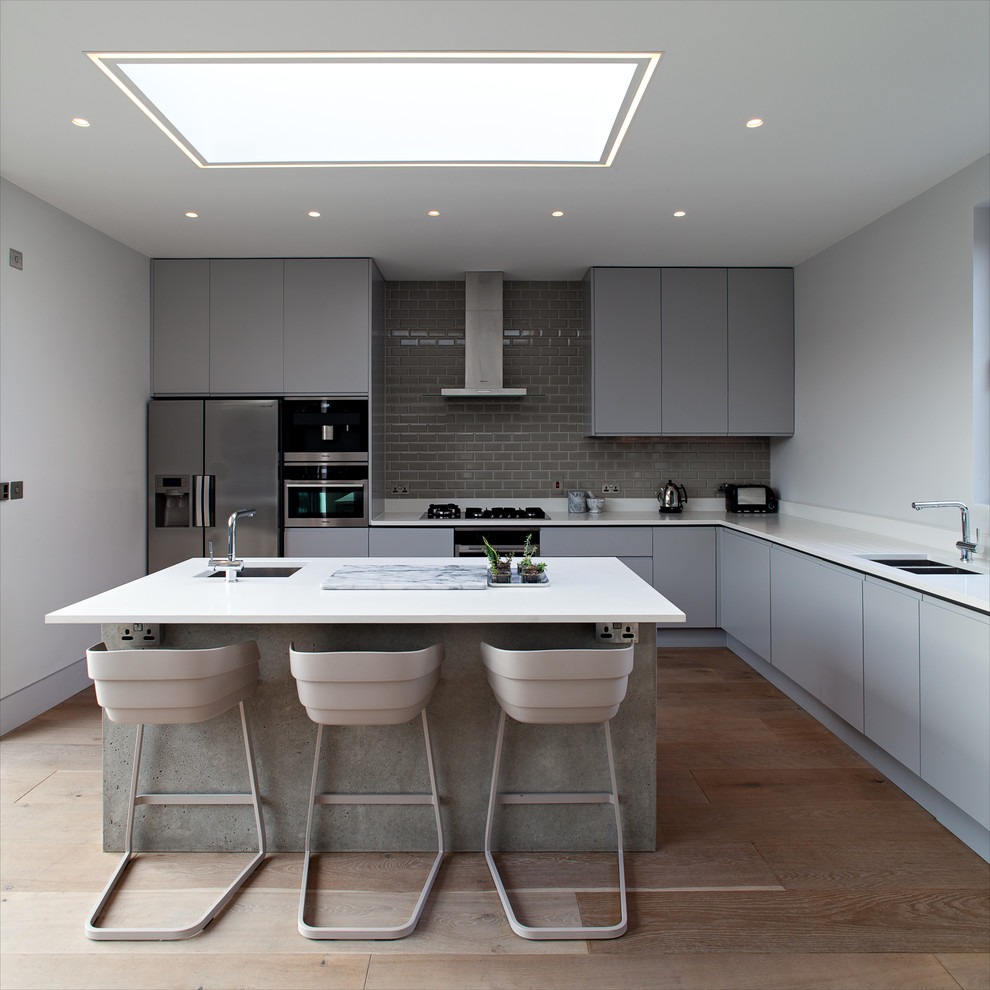 Example of a mid-sized trendy l-shaped medium tone wood floor open concept kitchen design in London with an undermount sink, flat-panel cabinets, gray cabinets, gray backsplash, subway tile backsplash, stainless steel appliances and an island