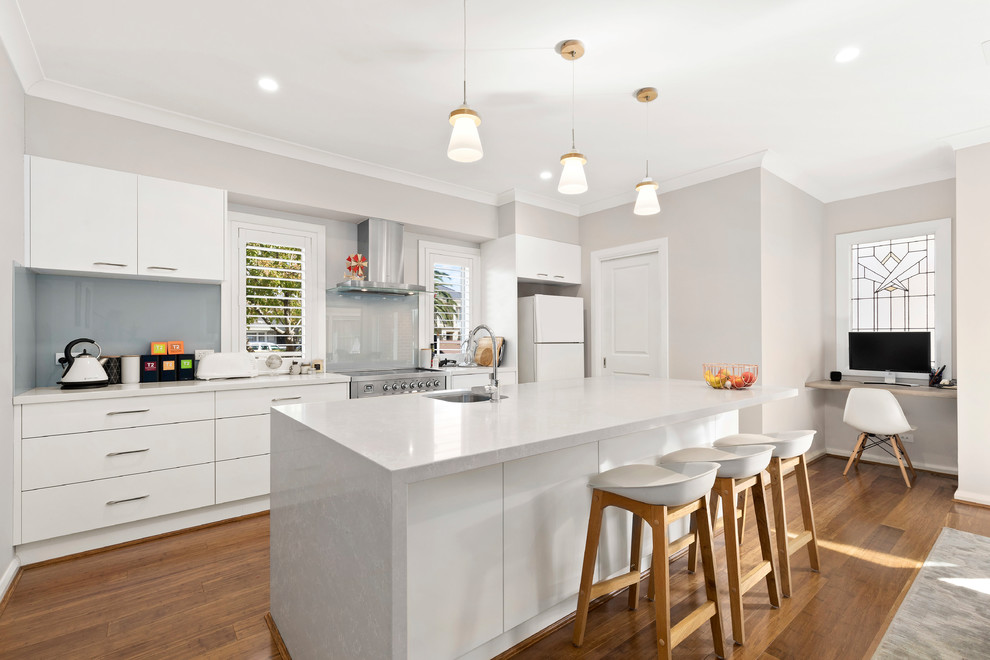 Photo of a medium sized victorian galley open plan kitchen in Melbourne with flat-panel cabinets, white cabinets, medium hardwood flooring, an island, brown floors, white worktops, a submerged sink, blue splashback, glass sheet splashback and stainless steel appliances.