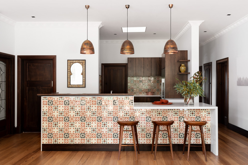 Offene Mediterrane Küche in U-Form mit flächenbündigen Schrankfronten, hellbraunen Holzschränken, bunter Rückwand, Küchengeräten aus Edelstahl, braunem Holzboden, Kücheninsel, braunem Boden und weißer Arbeitsplatte in Melbourne