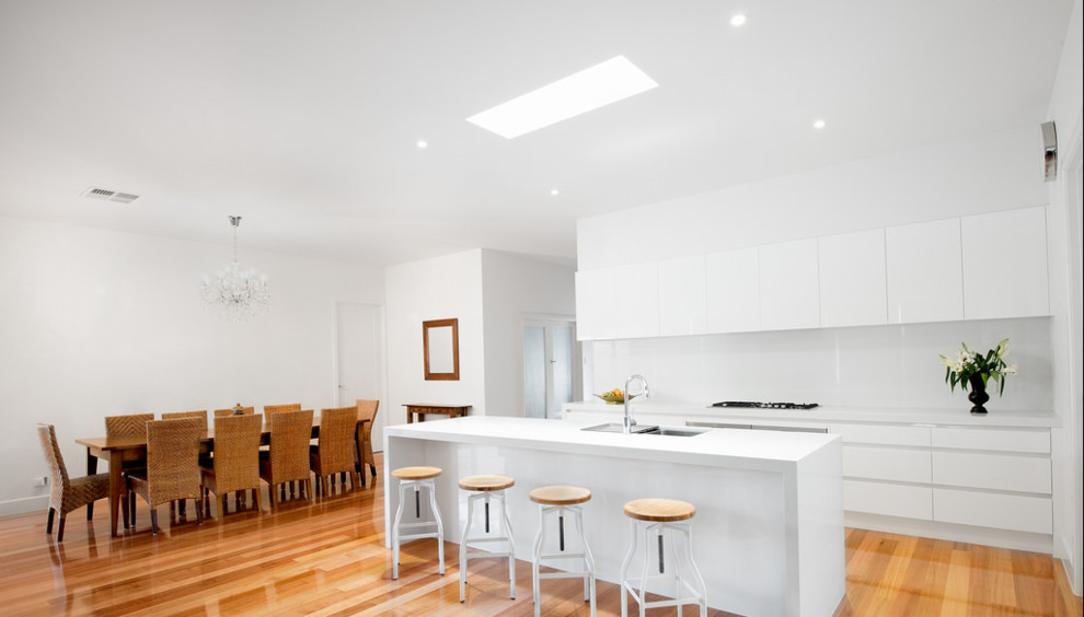 This is an example of a small contemporary single-wall kitchen/diner in Melbourne with a submerged sink, flat-panel cabinets, white cabinets, limestone worktops, white splashback, glass sheet splashback, stainless steel appliances, light hardwood flooring and an island.