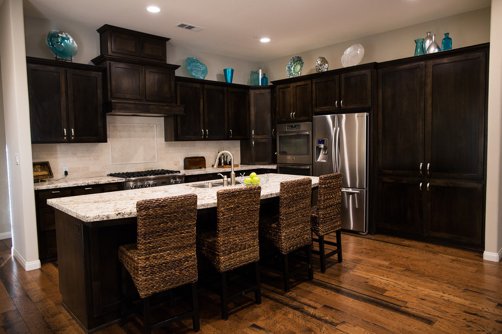 Trendy l-shaped medium tone wood floor open concept kitchen photo in Los Angeles with a double-bowl sink, shaker cabinets, dark wood cabinets, granite countertops, beige backsplash, stone tile backsplash, stainless steel appliances and an island