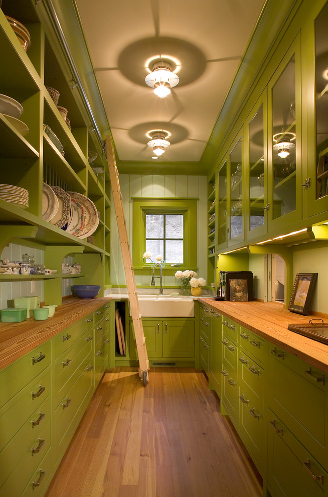 Inredning av ett lantligt kök, med en rustik diskho, luckor med glaspanel, gröna skåp, träbänkskiva, grönt stänkskydd, mellanmörkt trägolv och stänkskydd i glaskakel