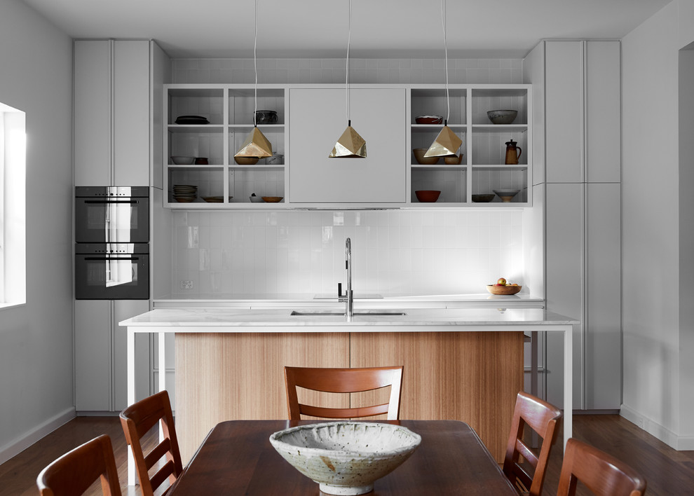 Imagen de cocina comedor moderna con armarios con paneles lisos, puertas de armario grises, salpicadero blanco, electrodomésticos negros, suelo de madera oscura, una isla y suelo marrón