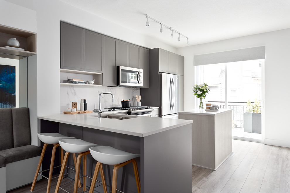 Photo of a scandinavian l-shaped open plan kitchen in Vancouver with a double-bowl sink, recessed-panel cabinets, grey cabinets, white splashback, stone slab splashback, stainless steel appliances, an island, white worktops and medium hardwood flooring.