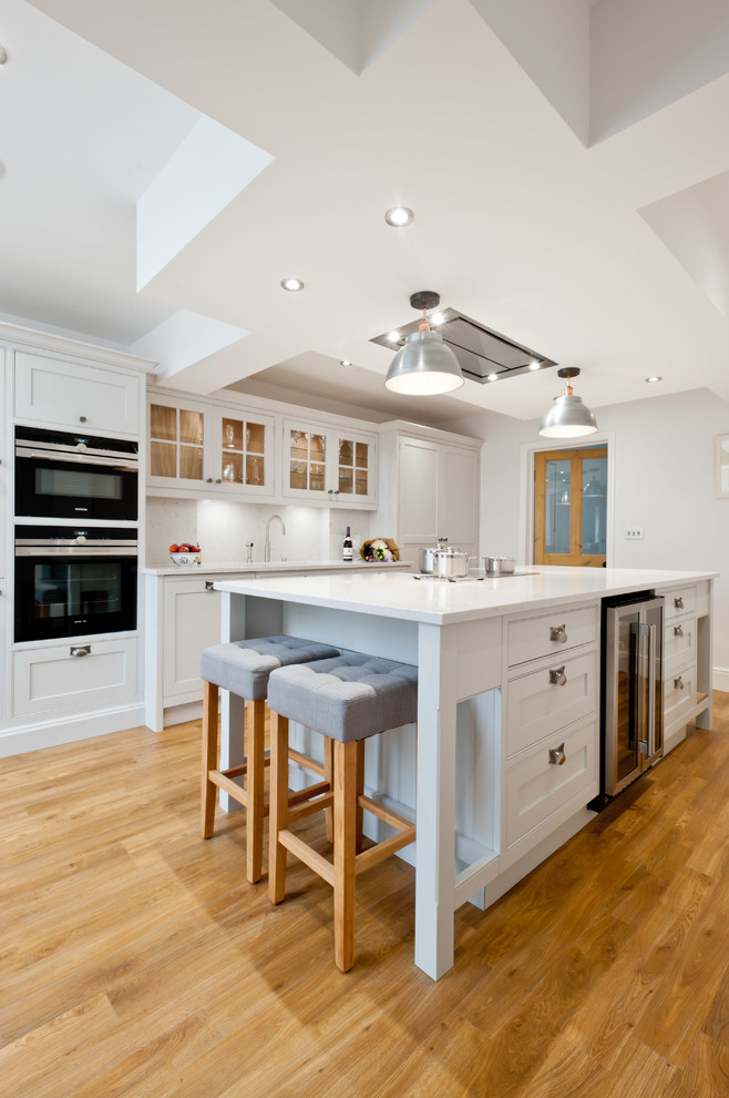 This is an example of a medium sized traditional galley kitchen in Kent with shaker cabinets, white cabinets, black appliances, light hardwood flooring, an island, beige floors and white worktops.