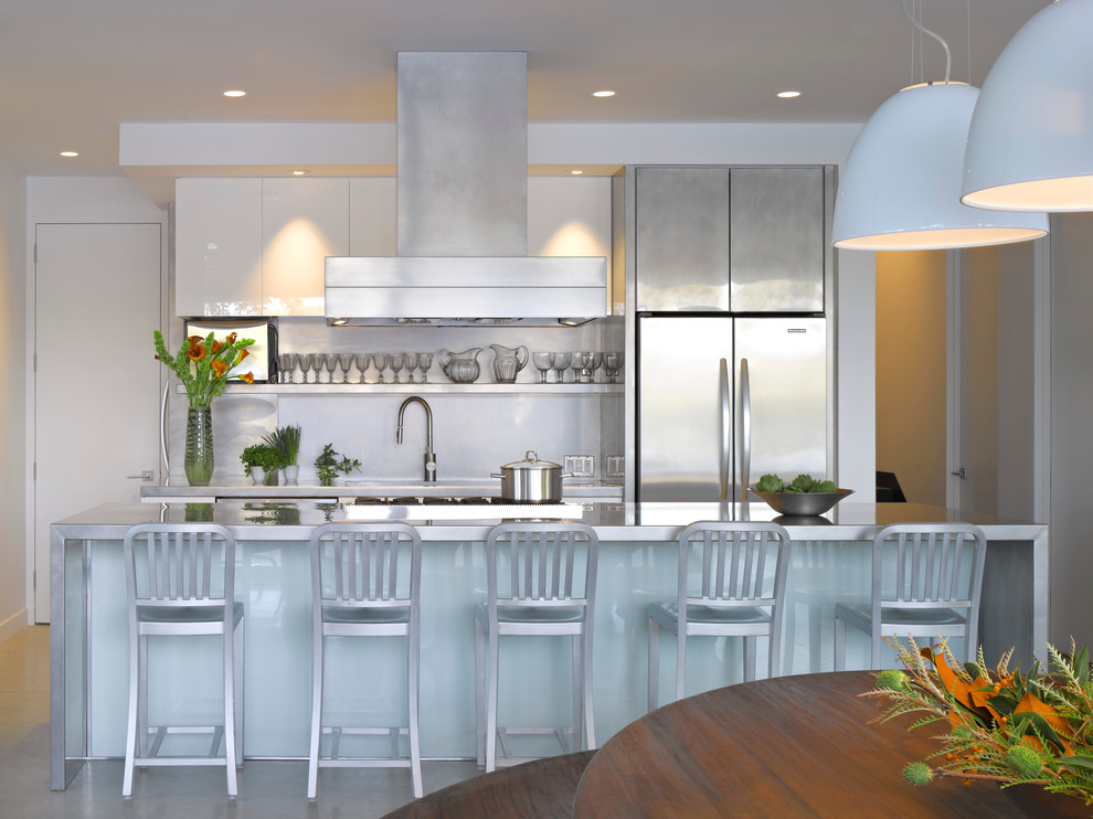 Example of a mid-sized trendy galley concrete floor and gray floor eat-in kitchen design in Burlington with stainless steel appliances, stainless steel countertops, flat-panel cabinets, an island, an undermount sink, white cabinets, white backsplash and stone slab backsplash