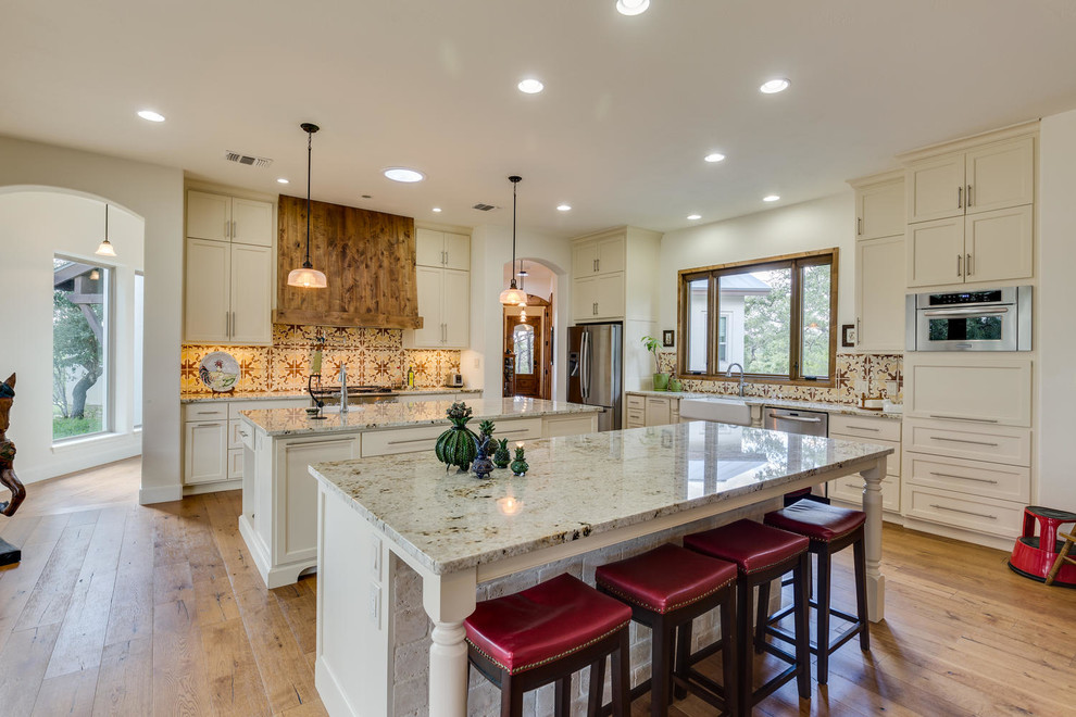 Photo of an expansive classic l-shaped open plan kitchen in Orange County with a built-in sink, shaker cabinets, white cabinets, engineered stone countertops, multi-coloured splashback, stainless steel appliances, light hardwood flooring, multiple islands, porcelain splashback and beige floors.