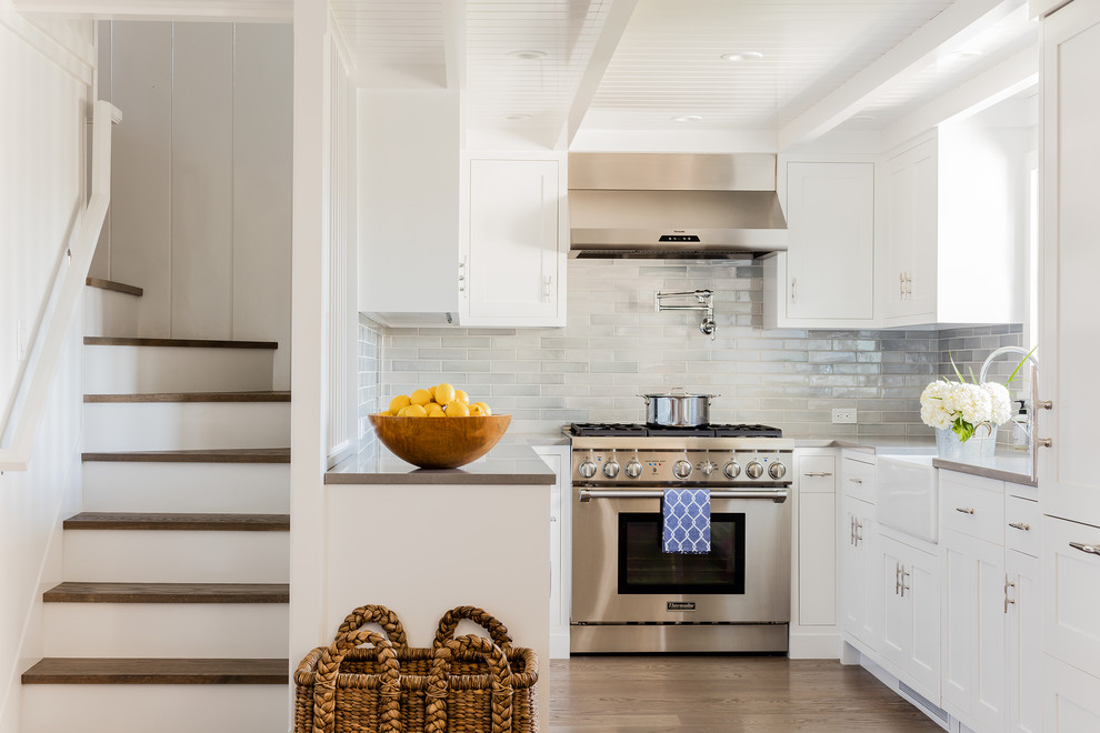 Photo of a medium sized beach style u-shaped kitchen/diner in Boston with a belfast sink, recessed-panel cabinets, white cabinets, composite countertops, grey splashback, glass tiled splashback, stainless steel appliances and light hardwood flooring.