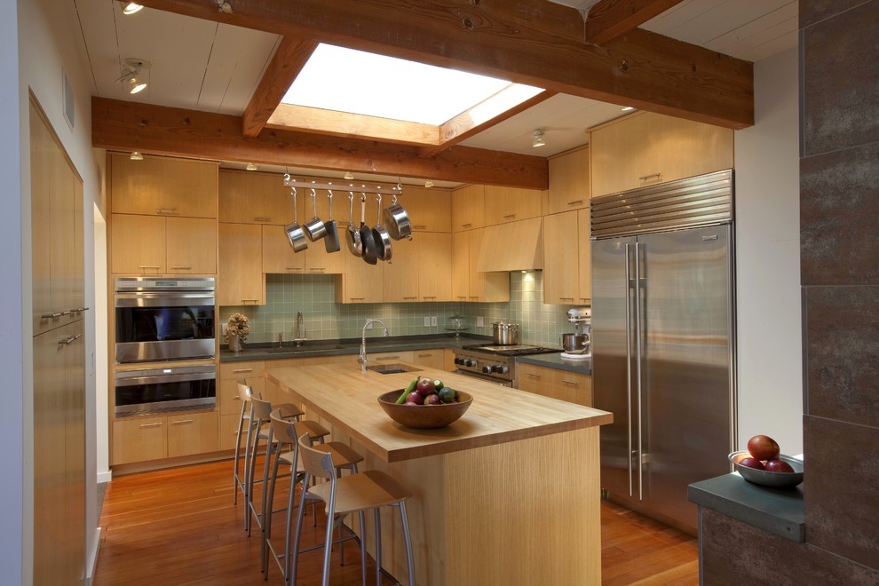 Foto på ett avskilt 50 tals l-kök, med släta luckor, skåp i mellenmörkt trä, träbänkskiva, grönt stänkskydd och rostfria vitvaror