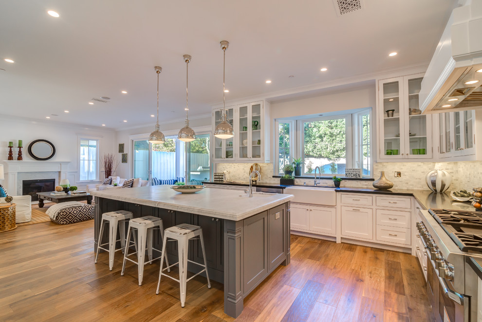 Example of a large transitional u-shaped medium tone wood floor eat-in kitchen design in Los Angeles with a farmhouse sink, glass-front cabinets, white cabinets, solid surface countertops, multicolored backsplash, porcelain backsplash, stainless steel appliances and an island