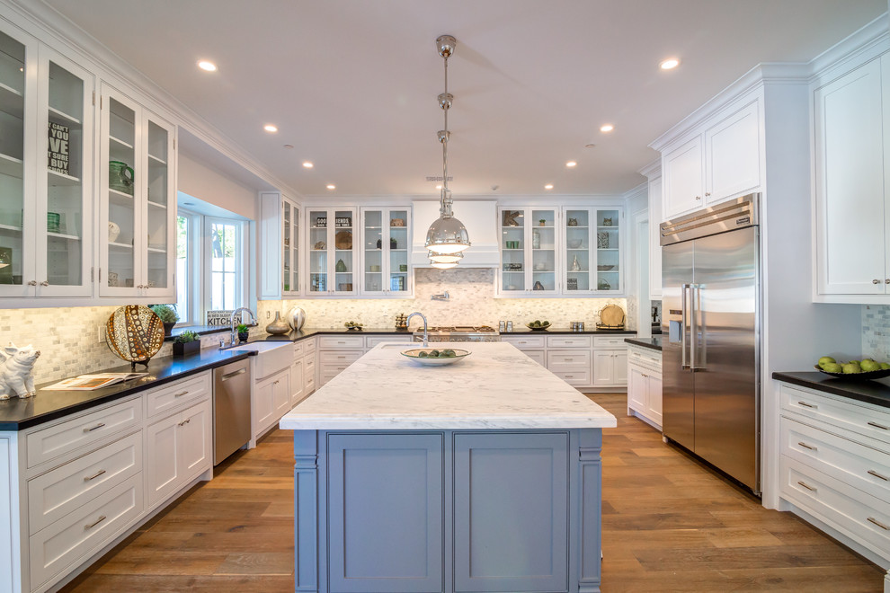 Example of a large transitional u-shaped medium tone wood floor eat-in kitchen design in Los Angeles with a farmhouse sink, glass-front cabinets, white cabinets, solid surface countertops, multicolored backsplash, porcelain backsplash, stainless steel appliances and an island