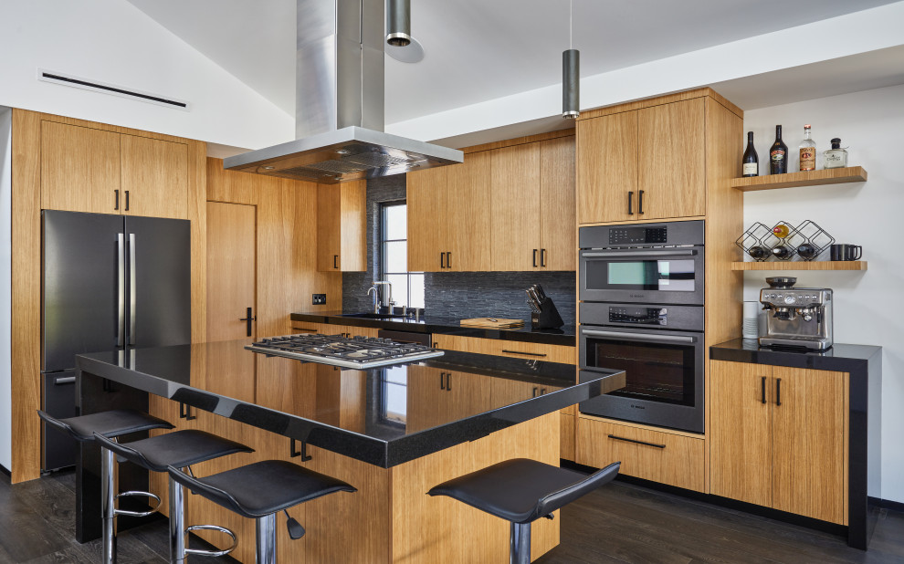 Moderne Küche in L-Form mit Unterbauwaschbecken, flächenbündigen Schrankfronten, hellbraunen Holzschränken, Küchenrückwand in Schwarz, schwarzen Elektrogeräten, dunklem Holzboden, Kücheninsel, schwarzem Boden und schwarzer Arbeitsplatte in Los Angeles
