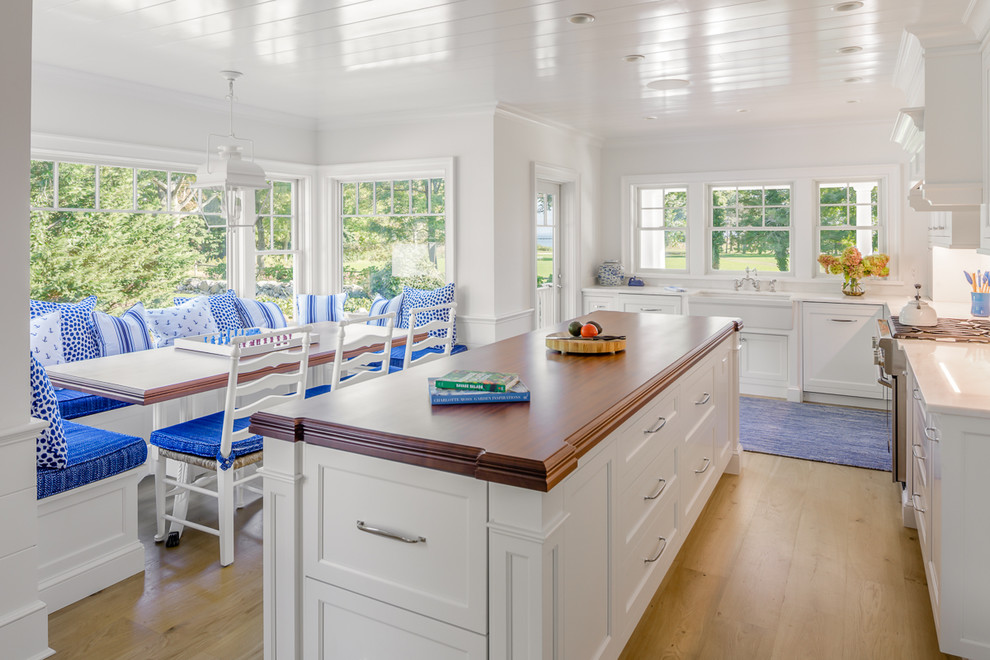 This is an example of a beach style l-shaped kitchen in Boston with a belfast sink, recessed-panel cabinets, white cabinets, wood worktops, stainless steel appliances, medium hardwood flooring, an island, brown floors and brown worktops.