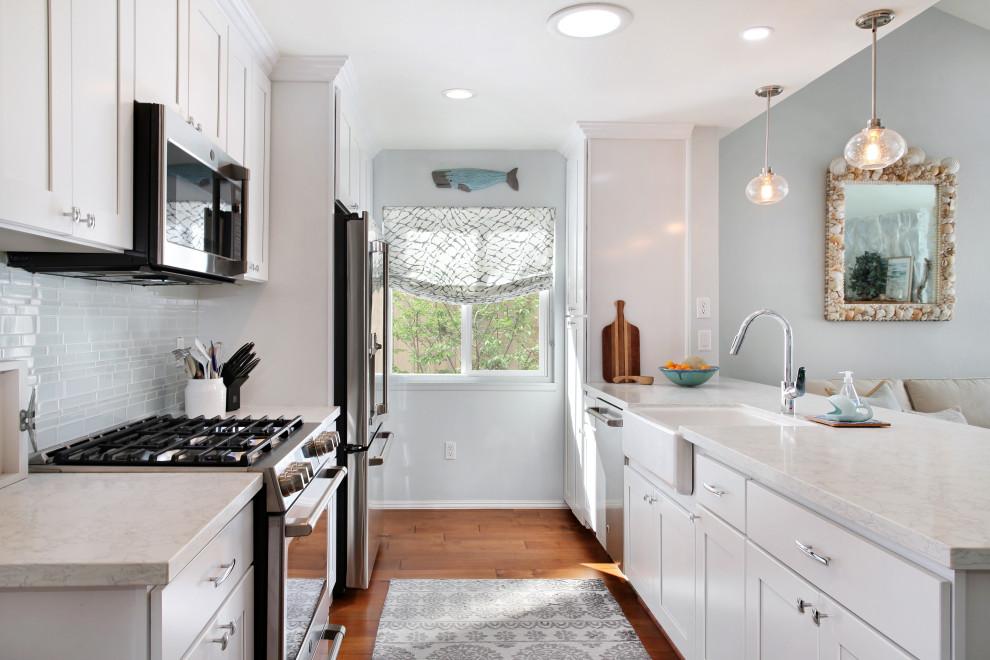 Photo of a small nautical galley kitchen in Orange County with a belfast sink, shaker cabinets, white cabinets, engineered stone countertops, blue splashback, glass tiled splashback, stainless steel appliances, medium hardwood flooring, brown floors and white worktops.