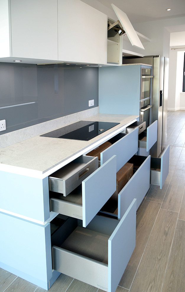 Foto på ett mellanstort funkis grå kök med öppen planlösning, med släta luckor, blå skåp, bänkskiva i kvarts, grått stänkskydd, glaspanel som stänkskydd och rostfria vitvaror