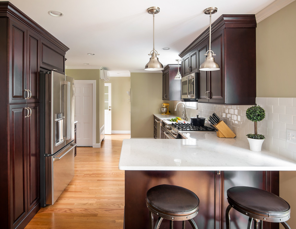 Inspiration for a mid-sized transitional l-shaped light wood floor eat-in kitchen remodel in Boston with an undermount sink, raised-panel cabinets, dark wood cabinets, quartz countertops, white backsplash, ceramic backsplash, stainless steel appliances, a peninsula and white countertops