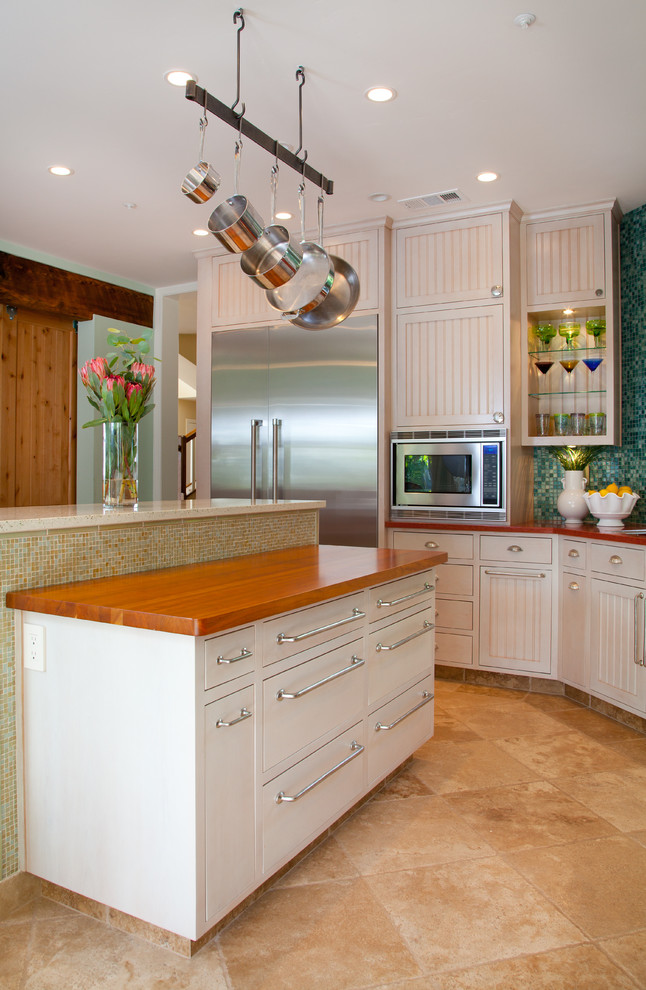 Foto på ett mellanstort tropiskt kök, med en undermonterad diskho, luckor med infälld panel, vita skåp, bänkskiva i återvunnet glas, grönt stänkskydd, rostfria vitvaror, kalkstensgolv och en köksö