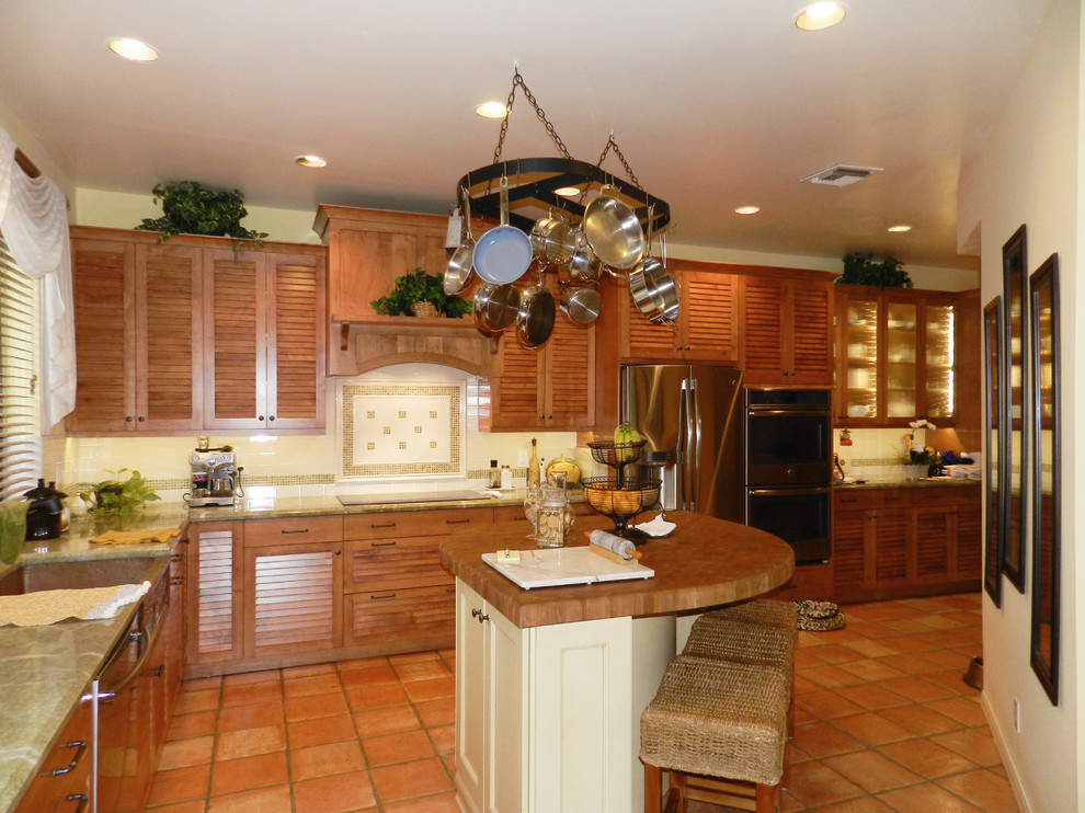 Große Küche in L-Form mit Landhausspüle, Lamellenschränken, hellbraunen Holzschränken, Küchenrückwand in Weiß, Rückwand aus Keramikfliesen, Küchengeräten aus Edelstahl, Terrakottaboden, Kücheninsel und braunem Boden in Miami