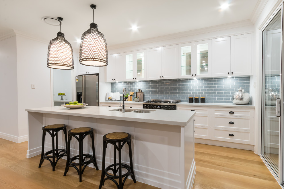 Beach style galley light wood floor kitchen photo in Townsville with a double-bowl sink, shaker cabinets, white cabinets, gray backsplash, subway tile backsplash, stainless steel appliances and an island