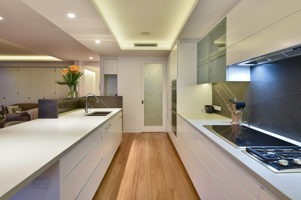 Photo of a large modern l-shaped open plan kitchen in Brisbane with a double-bowl sink, porcelain splashback, stainless steel appliances, an island, flat-panel cabinets, white cabinets, black splashback, light hardwood flooring and brown floors.