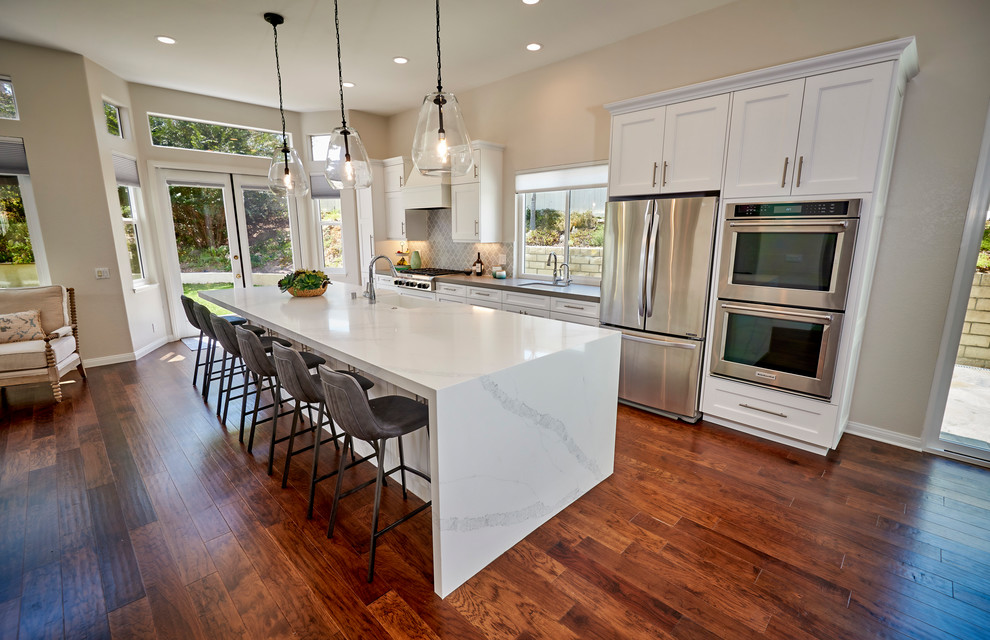 Example of a large transitional galley medium tone wood floor and beige floor open concept kitchen design in San Diego with an undermount sink, recessed-panel cabinets, white cabinets, quartz countertops, gray backsplash, marble backsplash, stainless steel appliances, an island and white countertops