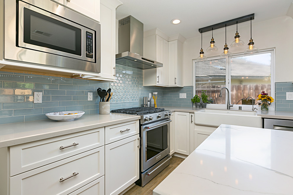 This is an example of an expansive coastal l-shaped kitchen/diner in San Diego with a belfast sink, recessed-panel cabinets, white cabinets, granite worktops, blue splashback, metro tiled splashback, stainless steel appliances, porcelain flooring, an island, brown floors, grey worktops and feature lighting.