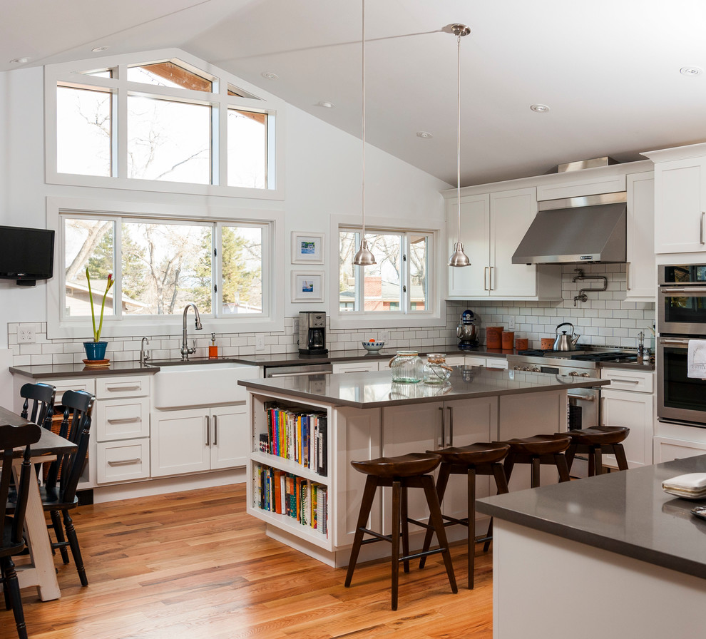 Design ideas for a large traditional u-shaped kitchen/diner in Denver with a belfast sink, shaker cabinets, engineered stone countertops, white splashback, metro tiled splashback, stainless steel appliances, medium hardwood flooring, an island, white cabinets and brown floors.