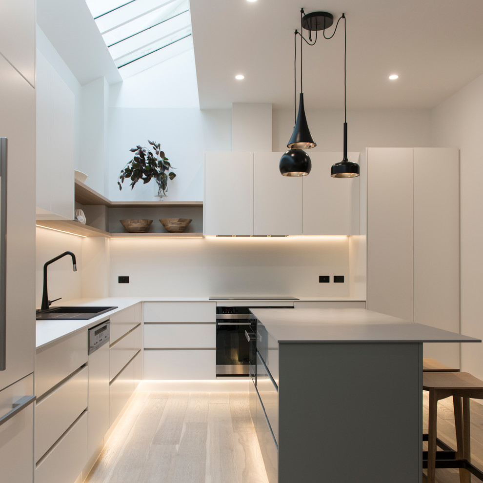 Photo of a medium sized contemporary l-shaped open plan kitchen in Melbourne with a built-in sink, white cabinets, composite countertops, white splashback, porcelain splashback, light hardwood flooring, an island, beige floors and grey worktops.