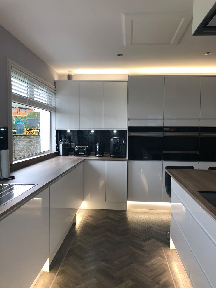 Large contemporary l-shaped kitchen in Glasgow with flat-panel cabinets, white cabinets, laminate countertops, black splashback, stainless steel appliances, vinyl flooring, an island, grey floors and brown worktops.