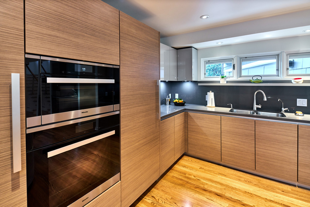 Aménagement d'une cuisine ouverte contemporaine en bois brun avec un placard à porte plane, un plan de travail en quartz modifié, une crédence grise, un électroménager noir, parquet clair et îlot.