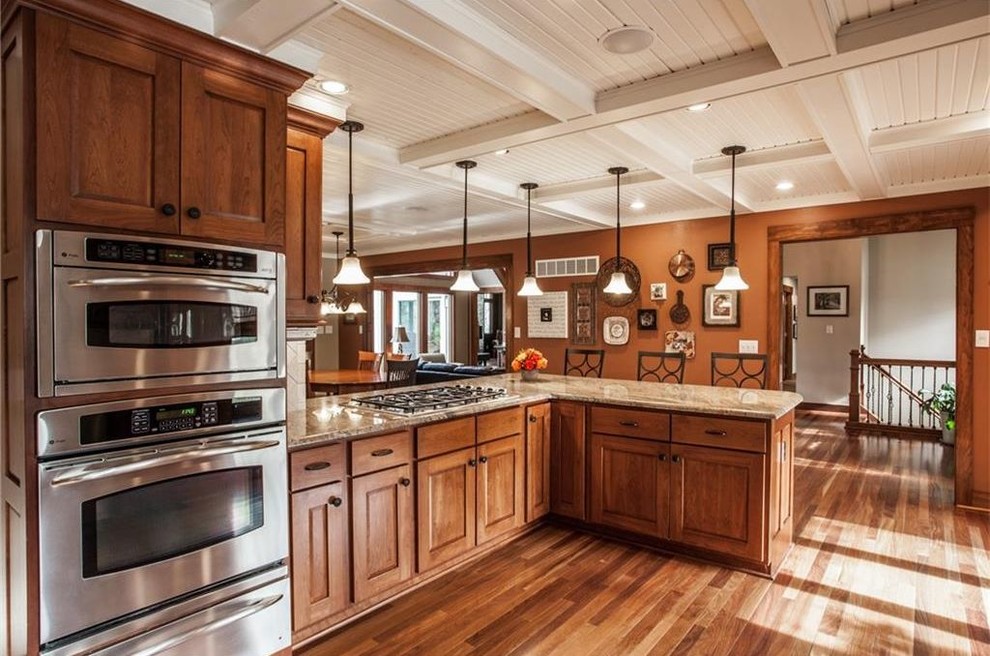 Photo of a large classic u-shaped open plan kitchen in Indianapolis with a submerged sink, recessed-panel cabinets, medium wood cabinets, granite worktops, beige splashback, ceramic splashback, stainless steel appliances, medium hardwood flooring, a breakfast bar, brown floors and beige worktops.