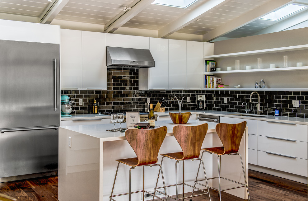 Photo of a large contemporary l-shaped kitchen/diner in Other with a submerged sink, flat-panel cabinets, white cabinets, black splashback, metro tiled splashback, stainless steel appliances, dark hardwood flooring, an island, engineered stone countertops and brown floors.