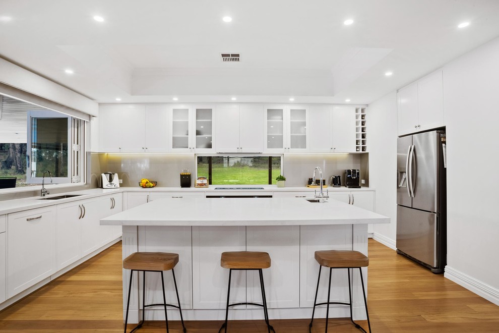 Klassisk inredning av ett stort vit vitt kök, med en köksö, en undermonterad diskho, vita skåp, svarta vitvaror, ljust trägolv, skåp i shakerstil, bänkskiva i koppar, stänkskydd med metallisk yta, glaspanel som stänkskydd och beiget golv