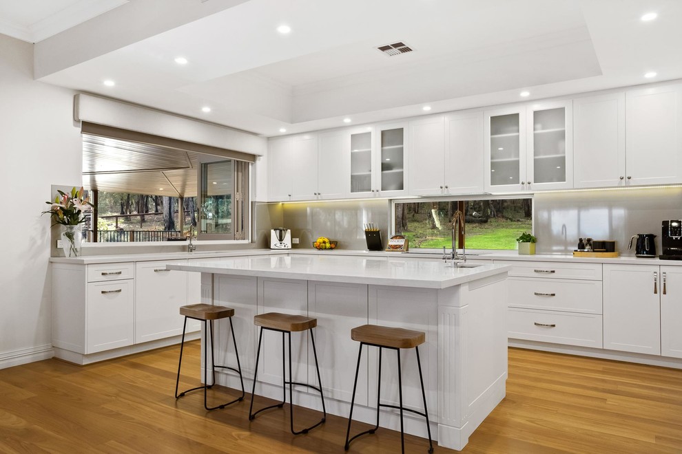 Large traditional galley kitchen/diner in Perth with an island, a submerged sink, white cabinets, black appliances, light hardwood flooring, white worktops, composite countertops, metallic splashback, glass sheet splashback, beige floors and flat-panel cabinets.