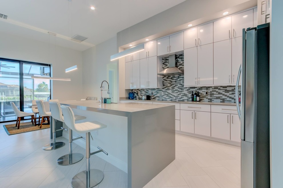 Inspiration for a large modern single-wall open plan kitchen in Other with a belfast sink, flat-panel cabinets, white cabinets, engineered stone countertops, multi-coloured splashback, glass tiled splashback, stainless steel appliances, an island, white floors and grey worktops.