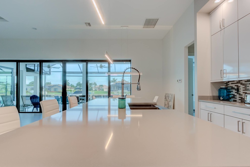 This is an example of a large modern single-wall open plan kitchen in Other with a belfast sink, flat-panel cabinets, white cabinets, engineered stone countertops, multi-coloured splashback, glass tiled splashback, stainless steel appliances, an island, white floors and grey worktops.