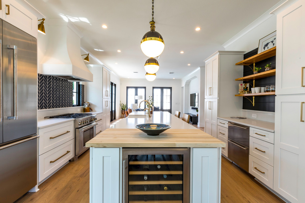Open concept kitchen - mid-sized transitional u-shaped light wood floor and brown floor open concept kitchen idea in Orlando with a farmhouse sink, flat-panel cabinets, white cabinets, black backsplash, porcelain backsplash, stainless steel appliances and an island