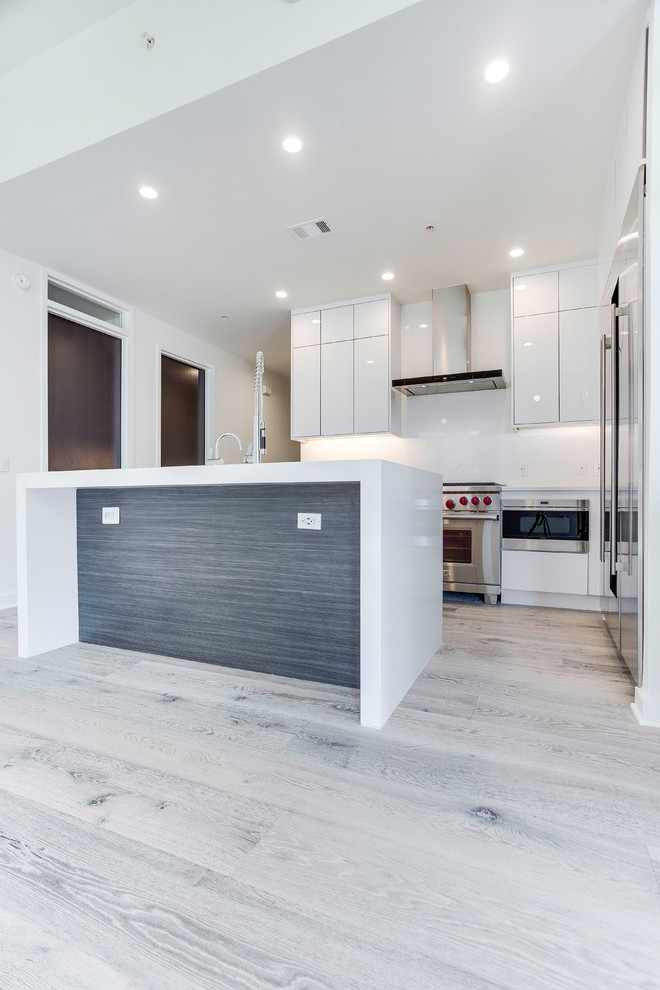Inspiration for a small modern kitchen in DC Metro with flat-panel cabinets, white cabinets, engineered stone countertops, white splashback, stainless steel appliances, light hardwood flooring, an island, grey floors and white worktops.