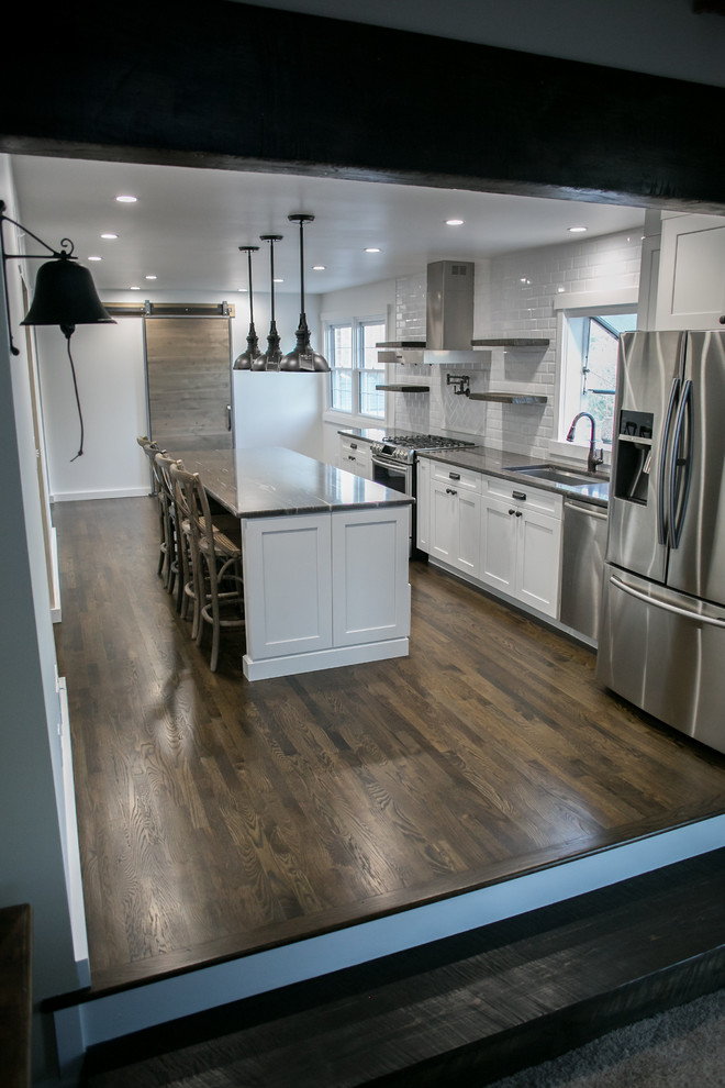 Exempel på ett mellanstort industriellt brun brunt kök, med en undermonterad diskho, skåp i shakerstil, vita skåp, granitbänkskiva, vitt stänkskydd, stänkskydd i tunnelbanekakel, rostfria vitvaror, mörkt trägolv, en köksö och brunt golv
