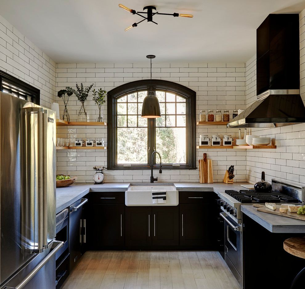 Mid-sized farmhouse u-shaped light wood floor and beige floor enclosed kitchen photo in New York with a farmhouse sink, black cabinets, concrete countertops, white backsplash, subway tile backsplash, stainless steel appliances, no island and flat-panel cabinets