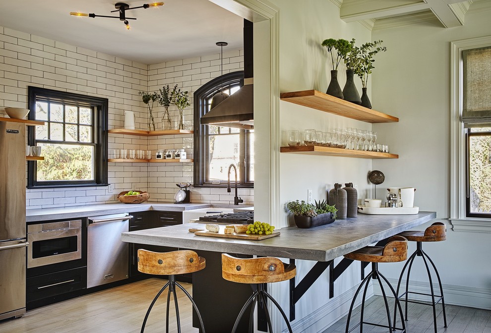 Inspiration for a mid-sized industrial u-shaped light wood floor and beige floor enclosed kitchen remodel in New York with a farmhouse sink, black cabinets, concrete countertops, white backsplash, subway tile backsplash, stainless steel appliances, no island and flat-panel cabinets