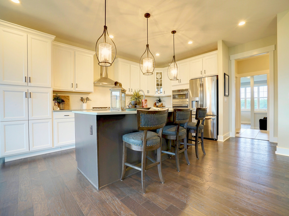 Example of a transitional single-wall dark wood floor and brown floor kitchen design in Cincinnati with a farmhouse sink, raised-panel cabinets, white cabinets, solid surface countertops, white backsplash, ceramic backsplash, stainless steel appliances and an island