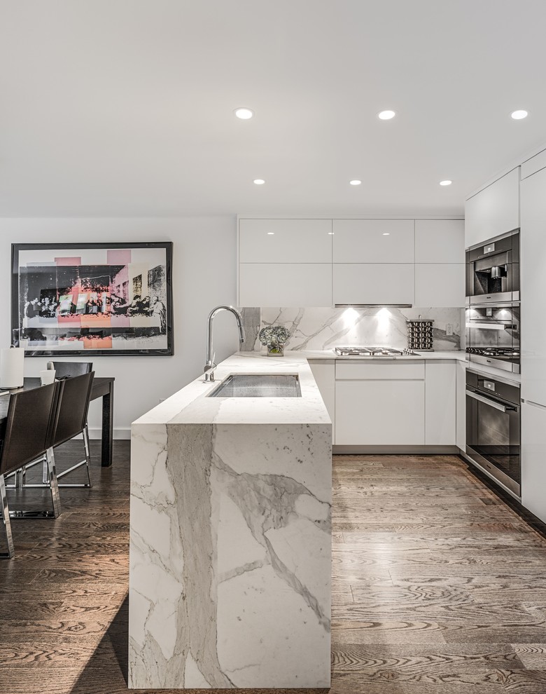 Photo of a medium sized contemporary u-shaped kitchen/diner in New York with a submerged sink, flat-panel cabinets, white cabinets, engineered stone countertops, white splashback, stone slab splashback, integrated appliances, dark hardwood flooring, white worktops, a breakfast bar and brown floors.