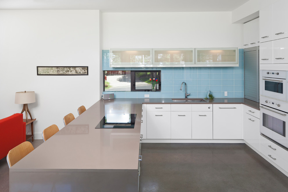 Inredning av ett modernt litet kök, med en undermonterad diskho, släta luckor, vita skåp, bänkskiva i kvartsit, blått stänkskydd, stänkskydd i glaskakel, vita vitvaror, betonggolv och en halv köksö