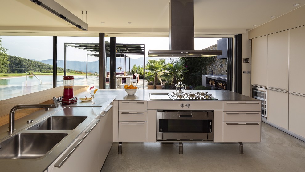 Modern inredning av ett avskilt, stort l-kök, med en dubbel diskho, släta luckor, vita skåp, bänkskiva i rostfritt stål, rostfria vitvaror, betonggolv och en halv köksö